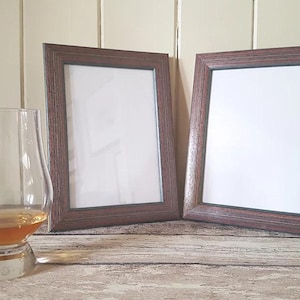 Puede incluir: Dos marcos de fotos de madera marrón con paspartú blanco y frentes de vidrio. Los marcos están sobre una superficie de madera. Un vaso transparente con un líquido marrón está en primer plano.