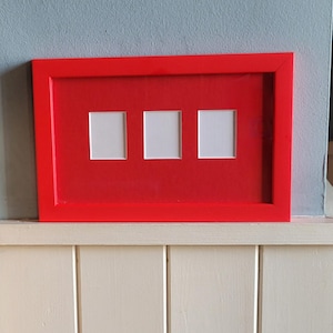 Könnte beinhalten: Ein leuchtend roter Bilderrahmen mit drei quadratischen Öffnungen. Der Rahmen hat ein klares, modernes Design, wahrscheinlich aus Holz. Die Öffnungen sind weiß und bilden einen starken Kontrast zum Rot.