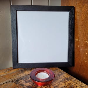 May include: A black framed picture with a white interior, resting on a wooden surface. A small, red glass candle holder with a lit white candle sits in front of the frame. The frame is rectangular.