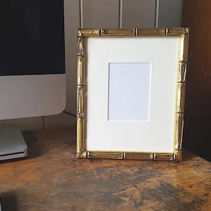 May include: A gold-colored picture frame with a white mat and a white background. The frame has a decorative design that resembles bamboo.