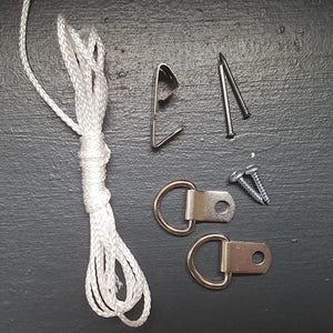 Può includere: Un assortimento di accessori per appendere quadri su sfondo scuro. Include un cordino bianco, un gancio metallico, due chiodi, due viti e due ganci ad anello a D.