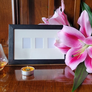 Peut inclure: Un cadre photo noir avec quatre ouvertures carrées blanches vides. Le cadre est posé sur une surface en bois avec une fleur de lys rose et une petite bougie allumée devant.
