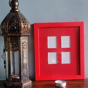 Può includere: Una lanterna decorativa in bronzo ornato con pannelli di vetro e una piccola candela all'interno. Una cornice rossa brillante con quattro aperture quadrate è accanto alla lanterna. Una piccola candela a lumino argentata è di fronte alla cornice.