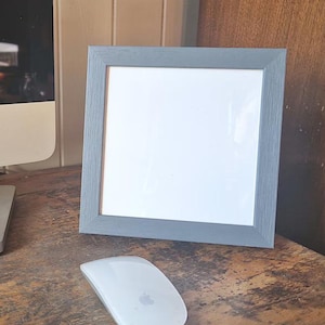 Peut inclure: Un cadre photo gris avec un passe-partout blanc et un fond blanc. Le cadre est posé sur une surface en bois.