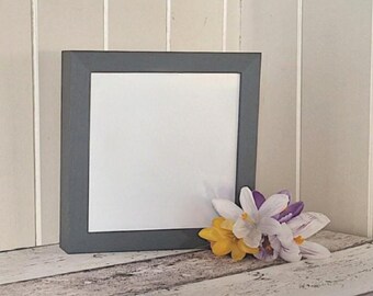 May include: A square, gray picture frame with a white interior, resting on a distressed wooden surface. A small bouquet of yellow, white, and purple flowers sits to the right of the frame. The background is a light beige.
