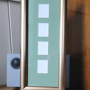 May include: A gold-coloured picture frame with a light green mount and four empty picture openings. The frame is standing on a wooden surface.