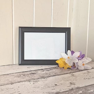 Pode incluir: Uma moldura retangular preta com interior branco, repousando sobre uma superfície de madeira envelhecida. Um pequeno buquê de flores amarelas, brancas e roxas é colocado à direita da moldura. O fundo apresenta painéis de madeira verticais de cor clara.