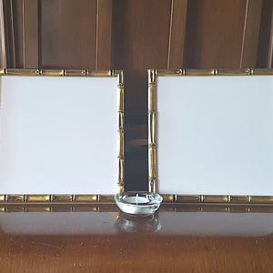 May include: Two gold bamboo picture frames with white mats, sitting on a wooden surface with a small glass candle holder in the center.