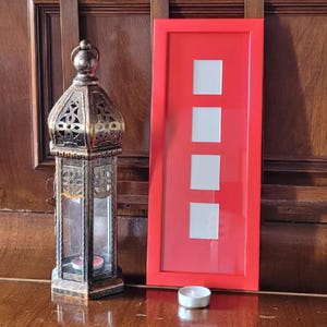 Può includere: Una lanterna decorativa in ottone con un involucro di vetro e una cornice rossa con quattro aperture quadrate bianche. Una piccola candela bianca è davanti alla cornice. Gli oggetti sono su una superficie di legno lucido.