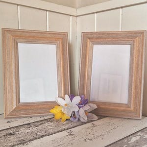 Puede incluir: Dos marcos de fotos de madera con un acabado marrón claro y paspartús blancos. Los marcos están vacíos y están sobre una superficie de madera blanqueada. Un grupo de crocus morados y blancos está delante de los marcos.