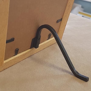 Pode incluir: Uma moldura de madeira com um fundo castanho e um cavalete de metal preto. A moldura é feita de madeira clara e tem um design simples e clássico. O suporte é curvo e fornece suporte para a moldura.