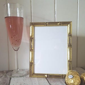 May include: A gold picture frame with a white mat and a glass of pink champagne in the foreground. The frame is on a white wooden surface.