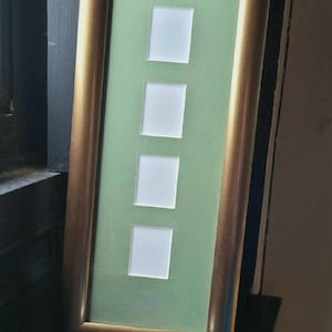 May include: A rectangular picture frame with a gold-coloured border. The frame holds a green mat with four square openings. A small, silver-coloured tea light candle sits in front of the frame.