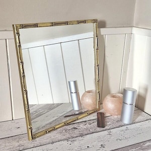 May include: A gold framed mirror with a bamboo design. The mirror is reflecting a white wall and two silver bottles.