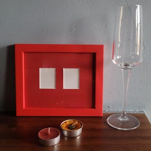 May include: A red picture frame with two openings, a champagne flute, and two small candles. One candle is red, and the other is in a silver container with orange and green decorations. The frame is on a wooden surface.