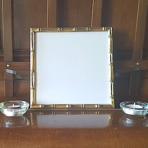 Op de afbeelding: Een gouden fotolijst met een bamboe-ontwerp staat op een houten oppervlak. Twee heldere glazen kaarshouders met witte kaarsen bevinden zich aan weerszijden van de lijst.