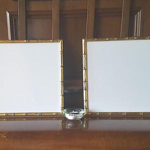 May include: Two gold-toned picture frames with white mats and a clear glass candle holder on a wooden surface.