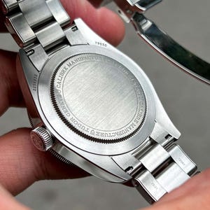 May include: Close-up of a stainless steel wristwatch. The watch back is visible, displaying the text "CALIBRE MANUFACTURE TUDOR GENEVE" around the edge. The serial number "2258952" is also visible. The watch band is made of stainless steel.