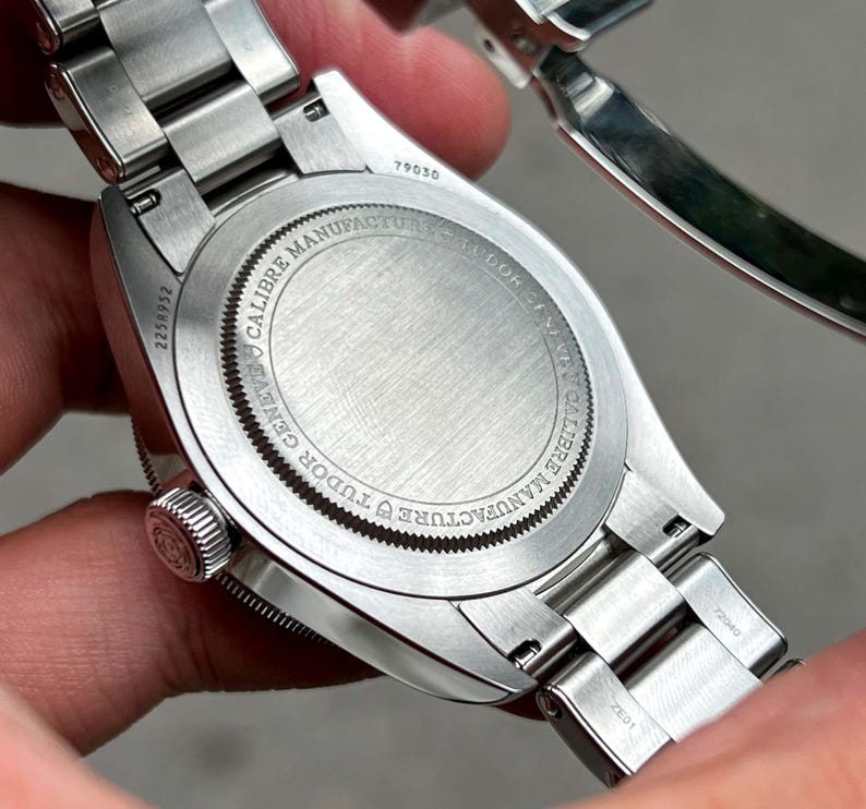 May include: Close-up of a stainless steel wristwatch. The watch back is visible, displaying the text "CALIBRE MANUFACTURE TUDOR GENEVE" around the edge. The serial number "2258952" is also visible. The watch band is made of stainless steel.
