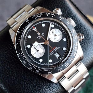 May include: A stainless steel Tudor Geneve chronograph watch with a black dial, white sub-dials, and a date window. The watch has a black bezel with white markings and a stainless steel bracelet. The watch is officially certified, Swiss made.
