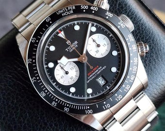 May include: A stainless steel Tudor Geneve chronograph watch with a black dial, white sub-dials, and a date window at the 6 o'clock position. The watch features a black bezel with white markings and a stainless steel bracelet. The watch is officially certified.