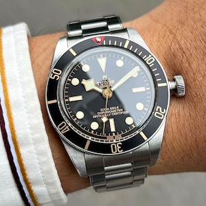 May include: A stainless steel wristwatch with a black dial and a black rotating bezel. The watch has luminous markers and hands, and the text "TUDOR GENEVE" is visible on the dial. The watch band is also stainless steel.