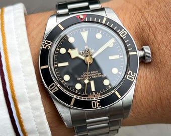 May include: A stainless steel wristwatch with a black dial and a black rotating bezel. The watch has luminous markers and hands, and the text "TUDOR GENEVE" is visible on the dial. The watch band is also stainless steel.