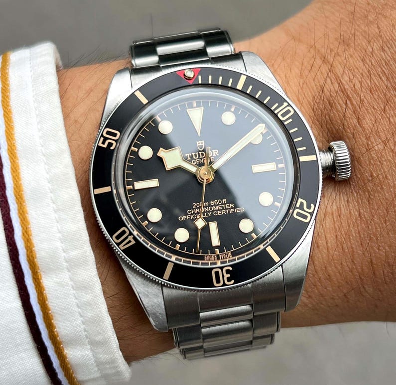 May include: A stainless steel wristwatch with a black dial and a black rotating bezel. The watch has luminous markers and hands, and the text "TUDOR GENEVE" is visible on the dial. The watch band is also stainless steel.