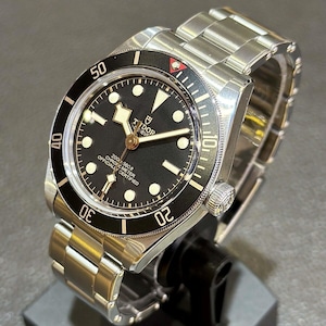 May include: A stainless steel wristwatch with a black dial and a black rotating bezel. The watch features luminous hour markers and hands, and the brand name "TUDOR" is displayed prominently on the dial. The watch is displayed on a black stand.