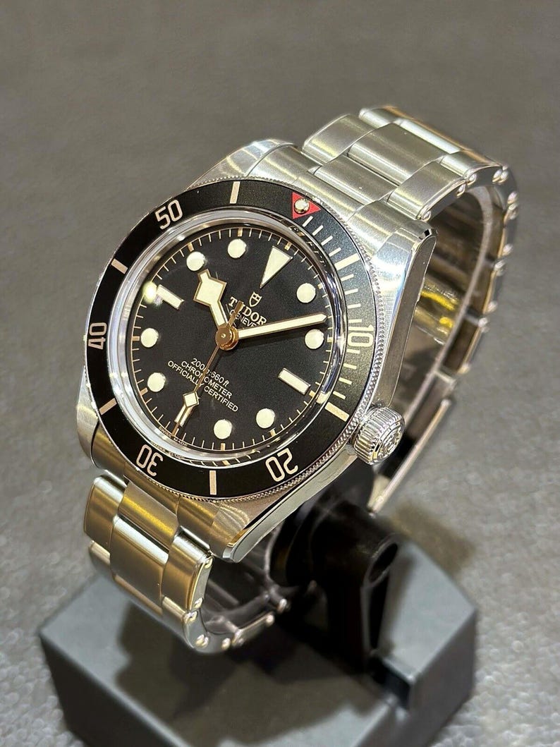 May include: A stainless steel wristwatch with a black dial and a black rotating bezel. The watch features luminous hour markers and hands, and the brand name "TUDOR" is displayed prominently on the dial. The watch is displayed on a black stand.