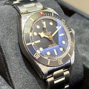 May include: A stainless steel wristwatch with a black dial and a silver-tone metal band. The watch face features luminous markers and hands, with the brand name "Tudor Geneve" visible. The bezel has a black insert with white numerals.