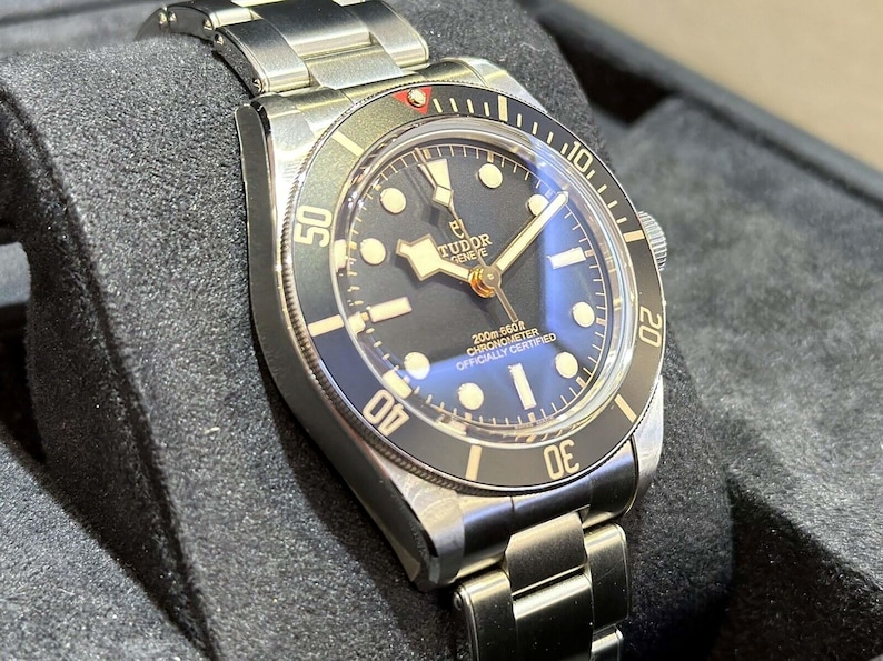 May include: A stainless steel wristwatch with a black dial and a silver-tone metal band. The watch face features luminous markers and hands, with the brand name "Tudor Geneve" visible. The bezel has a black insert with white numerals.