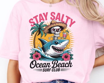 May include: Pink t-shirt with a graphic featuring a skeleton wearing a sombrero and sunglasses, riding a shark. The design includes the text "Stay Salty" and "Ocean Beach Surf Club."