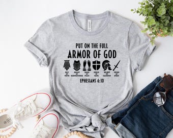 May include: A light gray t-shirt with the text "PUT ON THE FULL ARMOR OF GOD" and illustrations of armor pieces. Below the text are the words "BELT OF TRUTH", "BREAST OF RIGHTEOUSNESS", "SHOES OF PEACE", "SHIELD OF FAITH", "HELMET OF SALVATION", and "SWORD OF SPIRIT".