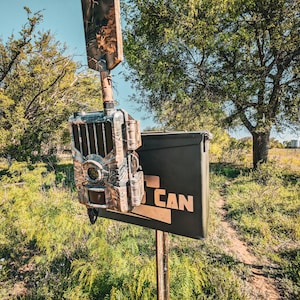 May include: A camouflage trail camera mounted on a wooden post with a black box that says "CAN" on it. The camera is in a wooded area with green grass and trees in the background.