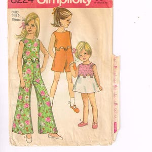 Może przedstawiać: Vintage wykrojnik krawiecki Simplicity 8224 dla odzieży dziecięcej. Wzór przedstawia trzy dziewczynki w różnych strojach: kombinezon w kwiaty, pomarańczowy top i szorty oraz różowo-białą sukienkę. Wykrojnik jest dla rozmiaru 6 z obwodem klatki piersiowej 63.5 cm.