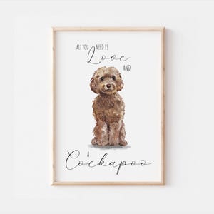 Può includere: Una stampa incorniciata con un'illustrazione ad acquerello di un cane Cockapoo. Il cane è marrone e soffice, seduto in posizione eretta. Il testo "All you need is Love and a Cockapoo" è scritto sopra e sotto il cane in elegante scrittura.