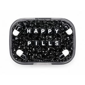 May include: A rectangular container with rounded corners filled with black rhinestones. The text HAPPY PILLS is spelled out in white letter beads across the center. Four black and white capsule-shaped objects are placed in the corners.