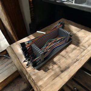 May include: A gray plastic organizer rack holding a collection of multicolored jumper wires. The rack features vertical slots that keep the ribbon cables separated, with black connector pins visible at both ends of the wire bundles.