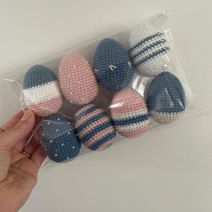 Op de afbeelding: Een set van acht gehaakte paaseieren in een doorzichtige plastic zak. De eieren zijn in verschillende patronen van blauw, roze en wit.