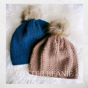 May include: Two knitted winter beanies with faux fur pom-poms. One beanie is a deep teal blue, the other is a light brown. The beanies are on a white, fluffy surface. The text "WINTER BEANIE" and "FRUENS HÆKLERIER" are visible.