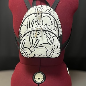 Könnte beinhalten: Ein kleiner weißer Rucksack mit schwarzen Hasen-Illustrationen und schwarzen Trägern. Der Rucksack hat zwei Reißverschluss-Zipper mit silbernen Anhängern.