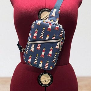 May include: A navy blue sling bag with a pattern of red and white lighthouses. The bag has a blue strap and a silver zipper. The bag is displayed on a maroon dress form.