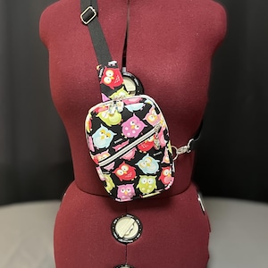 May include: A black crossbody bag with a colorful owl print. The bag has two zippered compartments and a silver chain strap.