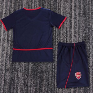 May include: A navy blue football jersey and shorts set with red trim. The shorts have the Arsenal logo on the right leg.