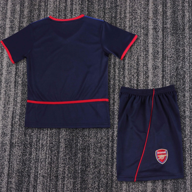May include: A navy blue football jersey and shorts set with red trim. The shorts have the Arsenal logo on the right leg.