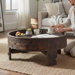 May include: A round, dark brown wooden coffee table with carved details and a low profile. The table holds a bowl of fruit, a succulent, two mugs, and an open book. The table is set on a woven rug in a living room setting.