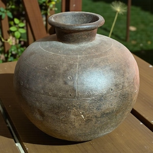 May include: A round, brown ceramic vase with a wide body and a narrow, flared rim. The vase has horizontal etched lines and small, evenly spaced circular indentations around its circumference. The surface has a mottled, aged appearance, suggesting a rustic or antique aesthetic.