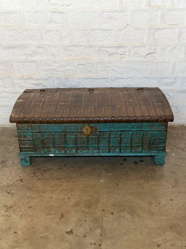 May include: A weathered wooden trunk with a curved top. The top is a dark brown color, while the body is a distressed teal. A brass-colored lock is centered on the front. The trunk is a decorative storage piece.