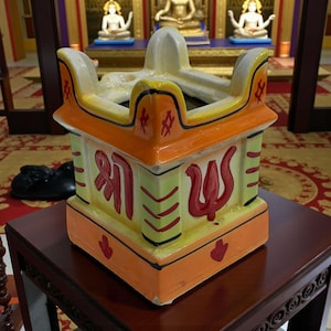May include: A colorful ceramic incense burner with a square base and a decorative top. The burner features orange, yellow, and green hues, with red script and symbols. It sits on a dark wooden table.